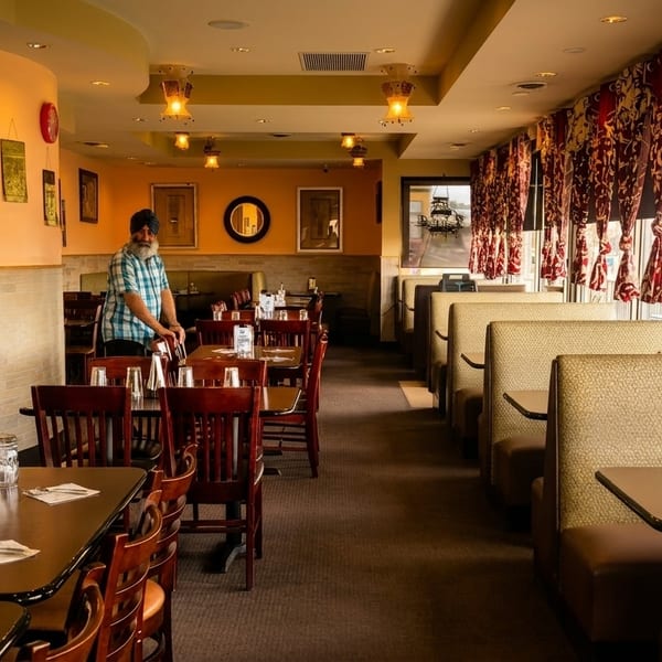 Restaurant interior with a man in a turban arranging chairs, featuring tables, booths, and patterned curtains.